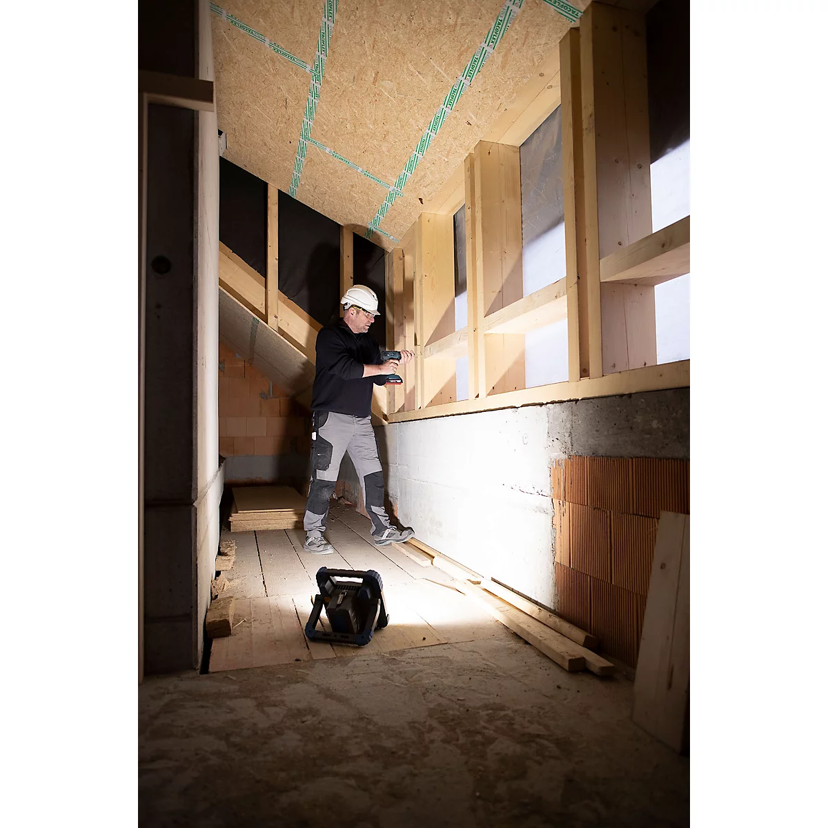 Ein Bauarbeiter mit Helm und Arbeitskleidung im Hausbau, hält eine Lampe. Im Vordergrund eine Baulampe, links der Durchgang.