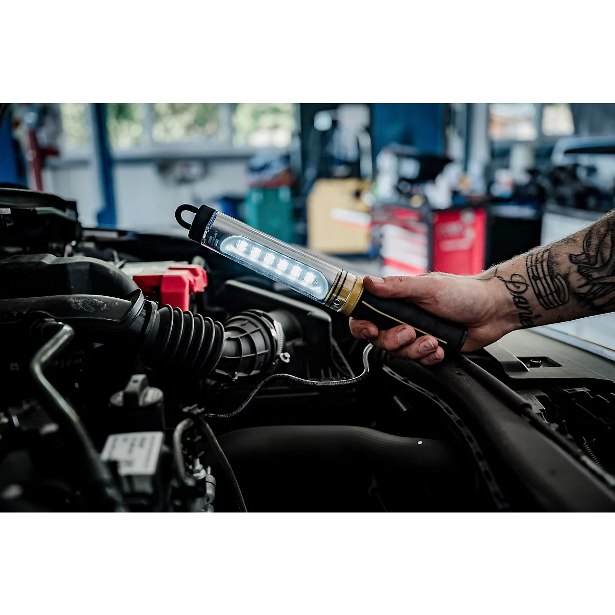 Une main tatouée tient une lampe LED brillante dans un compartiment moteur.