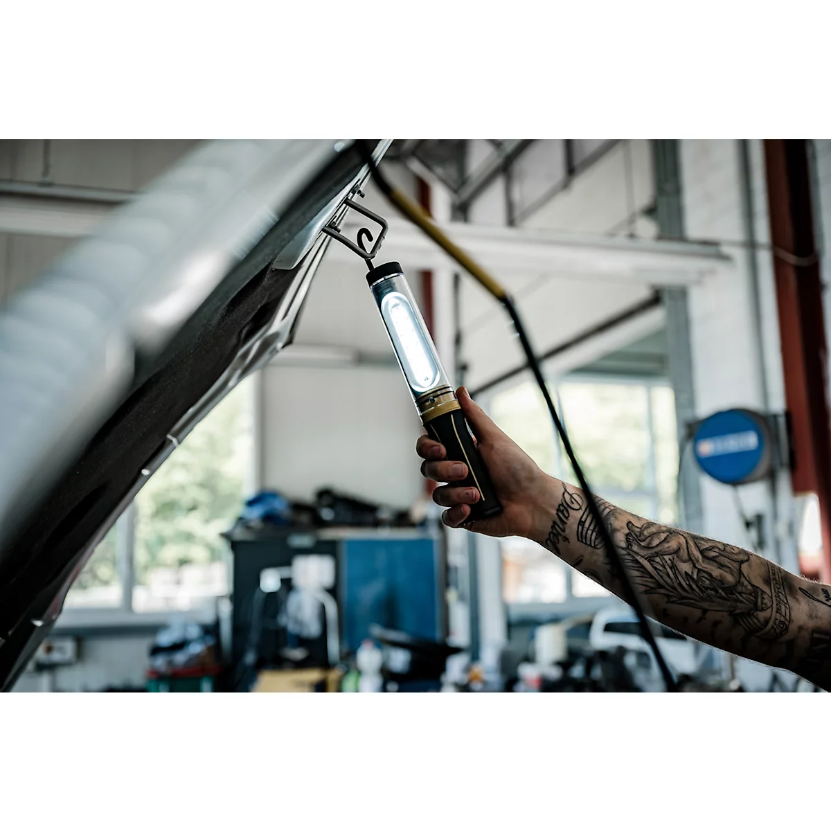 Une personne tient une lampe en forme de barre lumineuse sous le capot ouvert d'une voiture dans un atelier. Il y a un tatouage sur le bras.