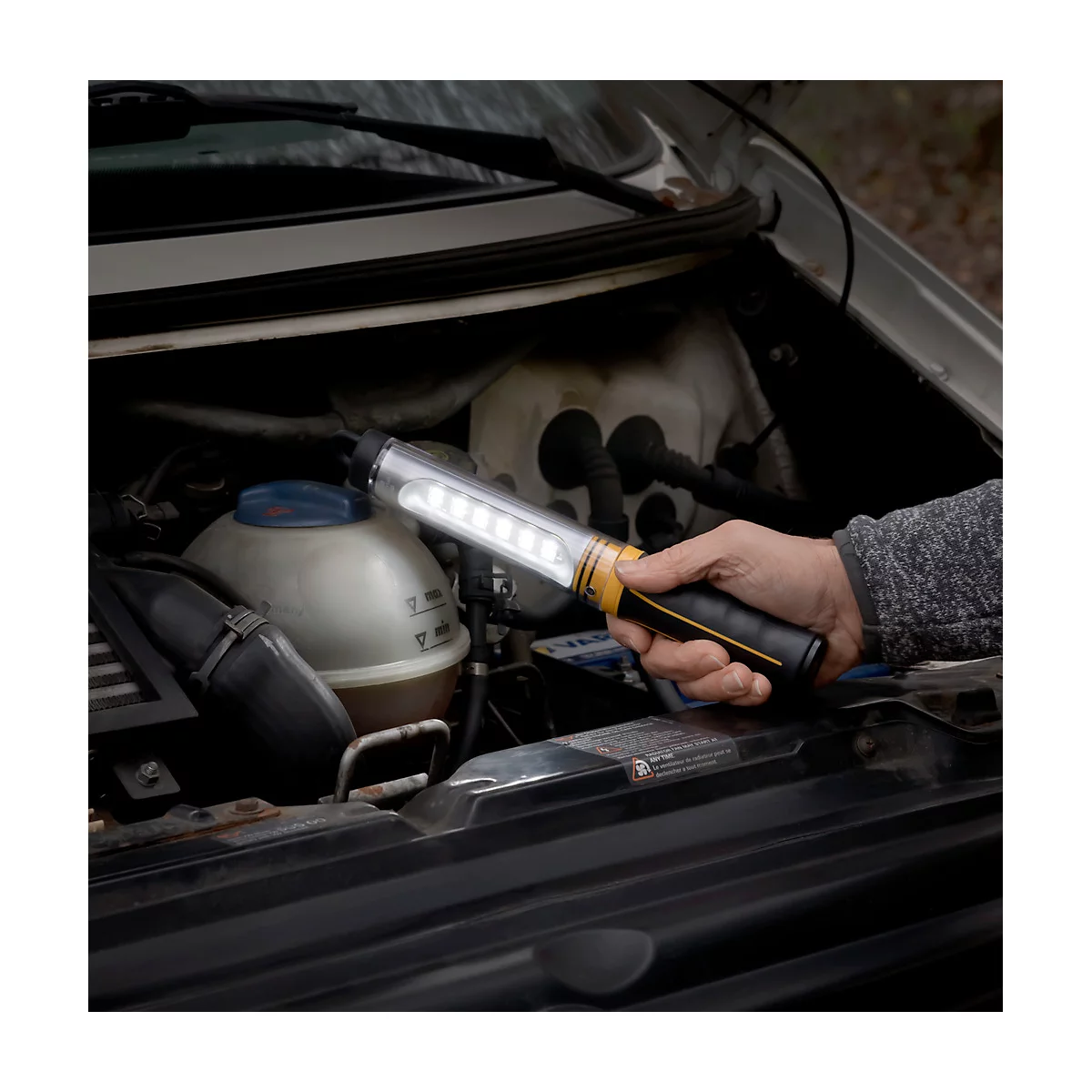 Une personne tient une lampe LED dans le compartiment moteur d'une voiture. La lampe est allumée. Le compartiment moteur est ouvert.