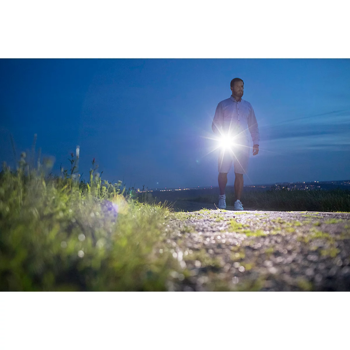 Un homme marche avec une lampe brillante la nuit. Herbe verte et chemin au premier plan, ville en arrière-plan.