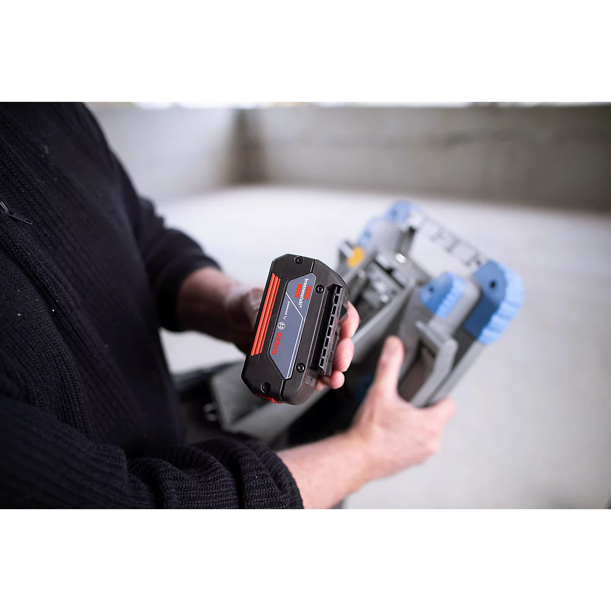 Homme tenant une batterie d'outil électrique et un étui. Batterie noire-rouge-bleue, étui gris-bleu. Arrière-plan flou.