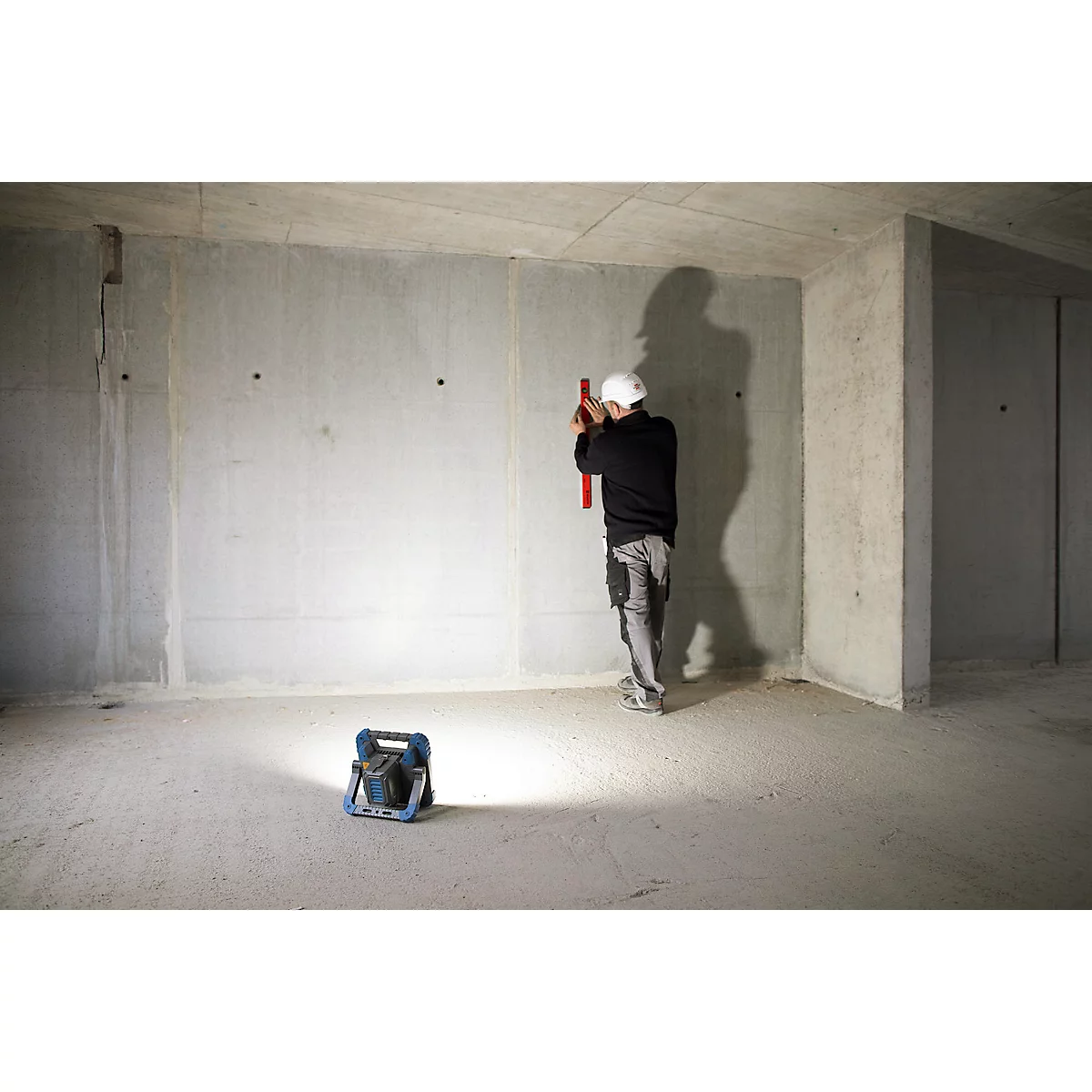 Un ouvrier du bâtiment mesure le mur avec un niveau à bulle dans une construction brute. Au premier plan, une lampe de chantier.