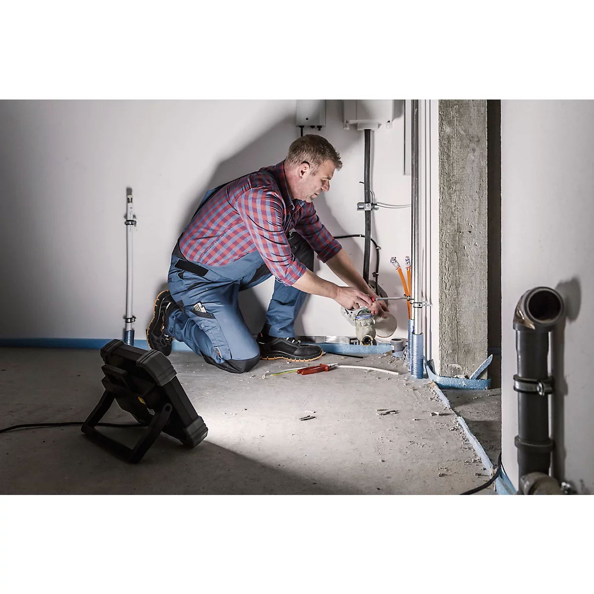 Un homme s'agenouille sur le sol, travaillant sur des tuyaux. Il porte des vêtements de travail, une lampe frontale éclaire le sol.