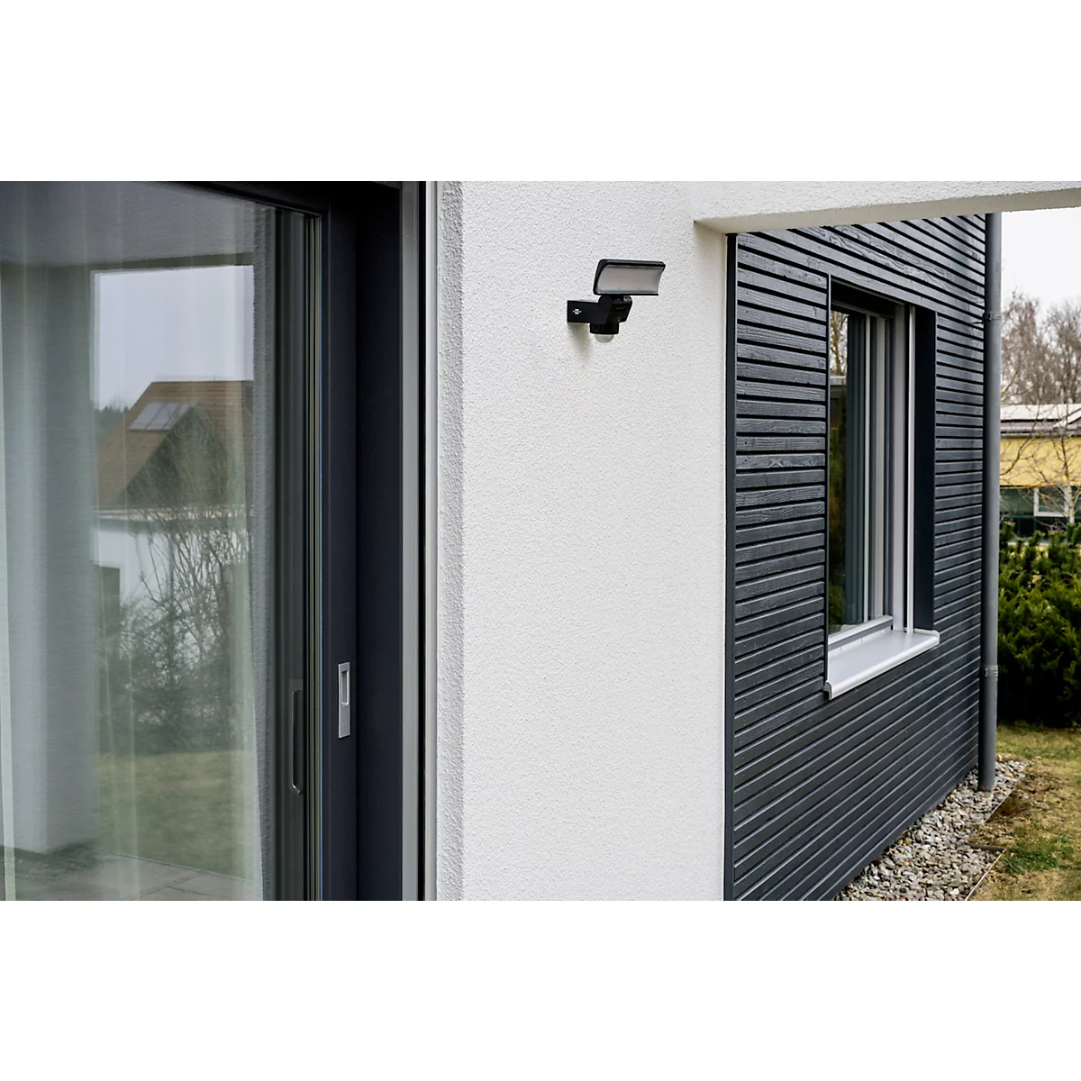 Buiten aan een muur van een huis staat een zwarte bewegingsmelder en een buitenlamp. Rechts een raam en grijze houten panelen.