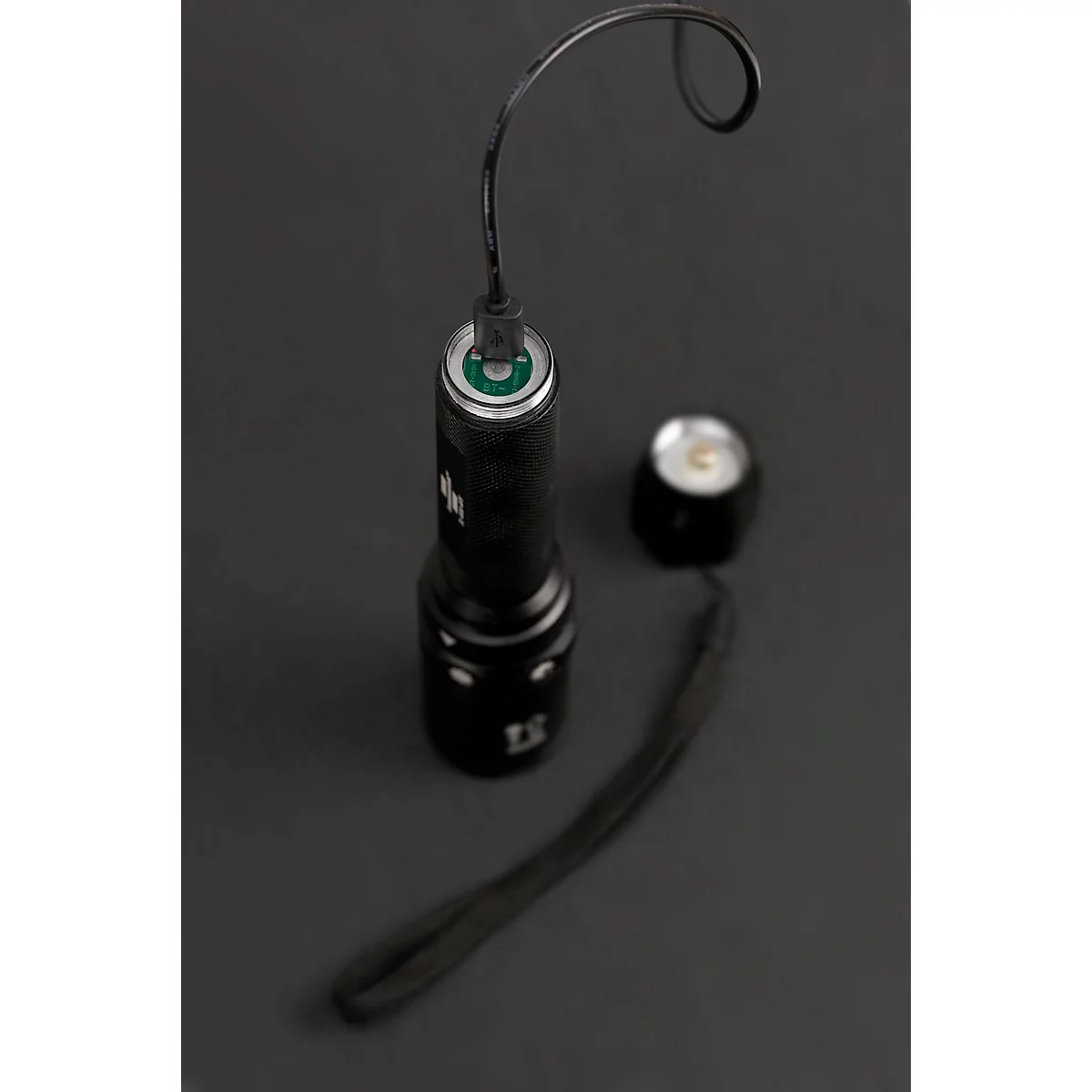 Schwarze Taschenlampe mit abgenommenem Kopf, dazu ein Handgelenksriemen. Auf dem grauen Hintergrund.