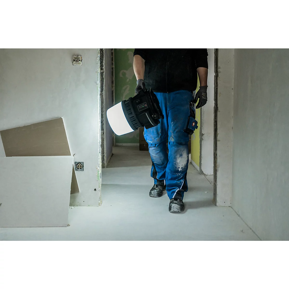 Persoon draagt lamp door gang in ruwbouw, blauwe werkkleding.