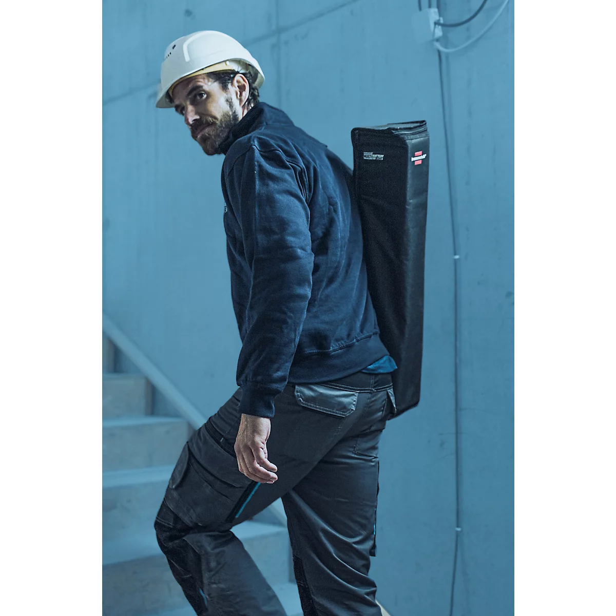 Un ouvrier du bâtiment en vêtements de travail et un casque porte un sac noir sur son épaule en montant un escalier.