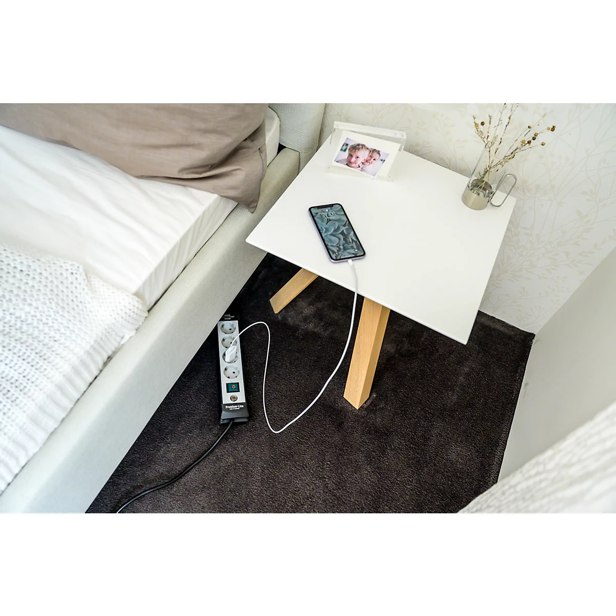 Un smartphone se charge sur une table de chevet blanche, à côté une multiprise. Sur la table se trouvent un petit vase avec des branches et une photo.