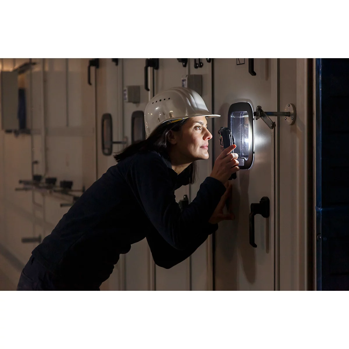 Een vrouw met een helm onderzoekt een schakelpaneel met een lamp. Ze lijkt geconcentreerd.