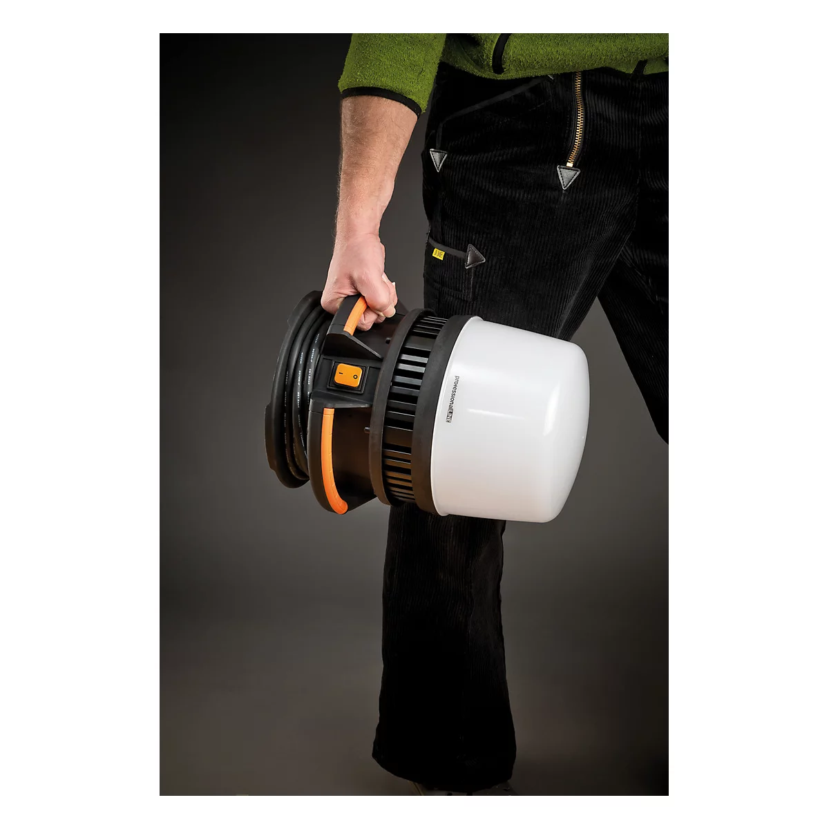Een persoon draagt een mobiele bouwplaatslamp, bestaande uit een witte lampenkap en een zwarte behuizing. De handgreep is oranje.