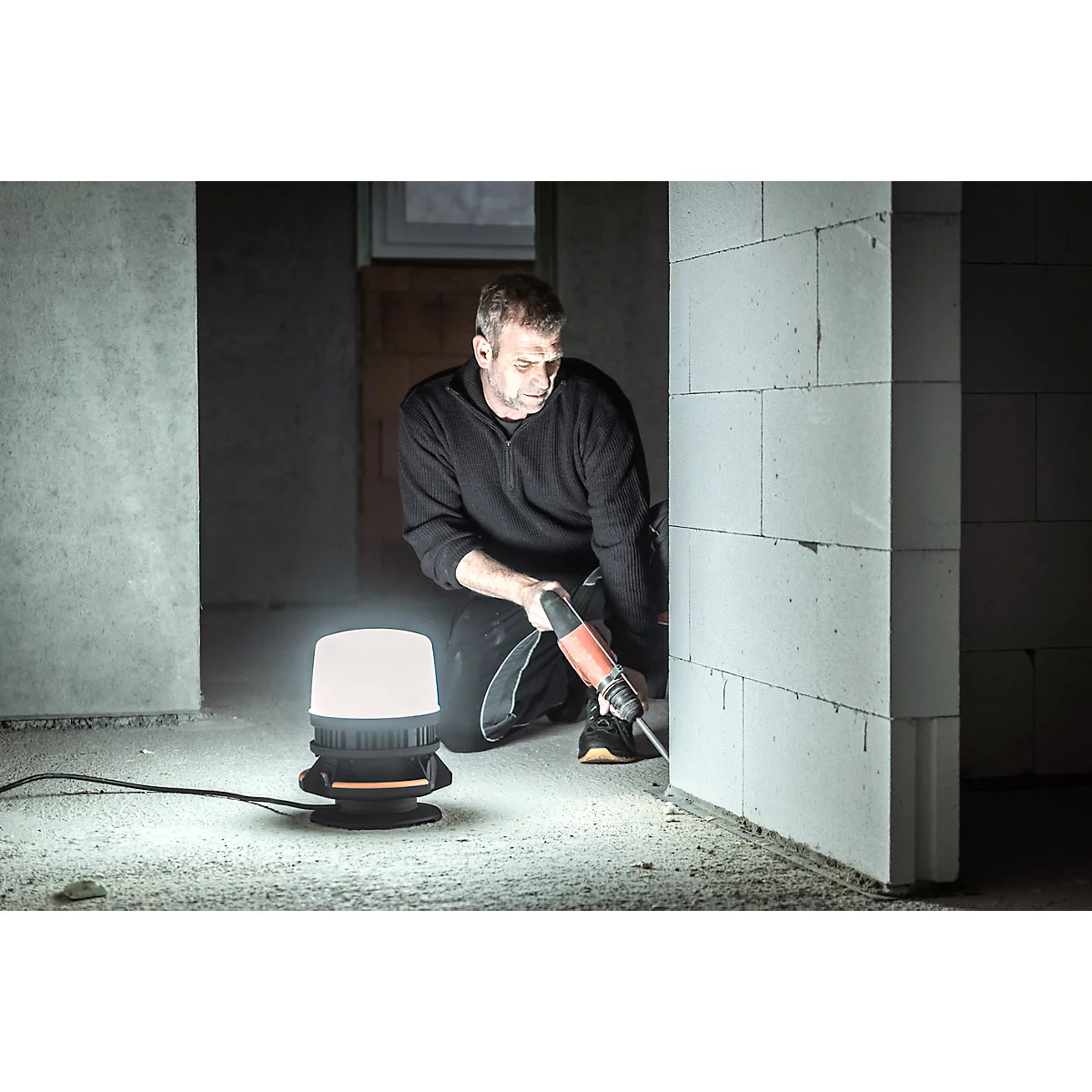 Een man knielt op een bouwplaats en gebruikt een slijpmachine op een muur, terwijl een heldere lamp naast hem schijnt.