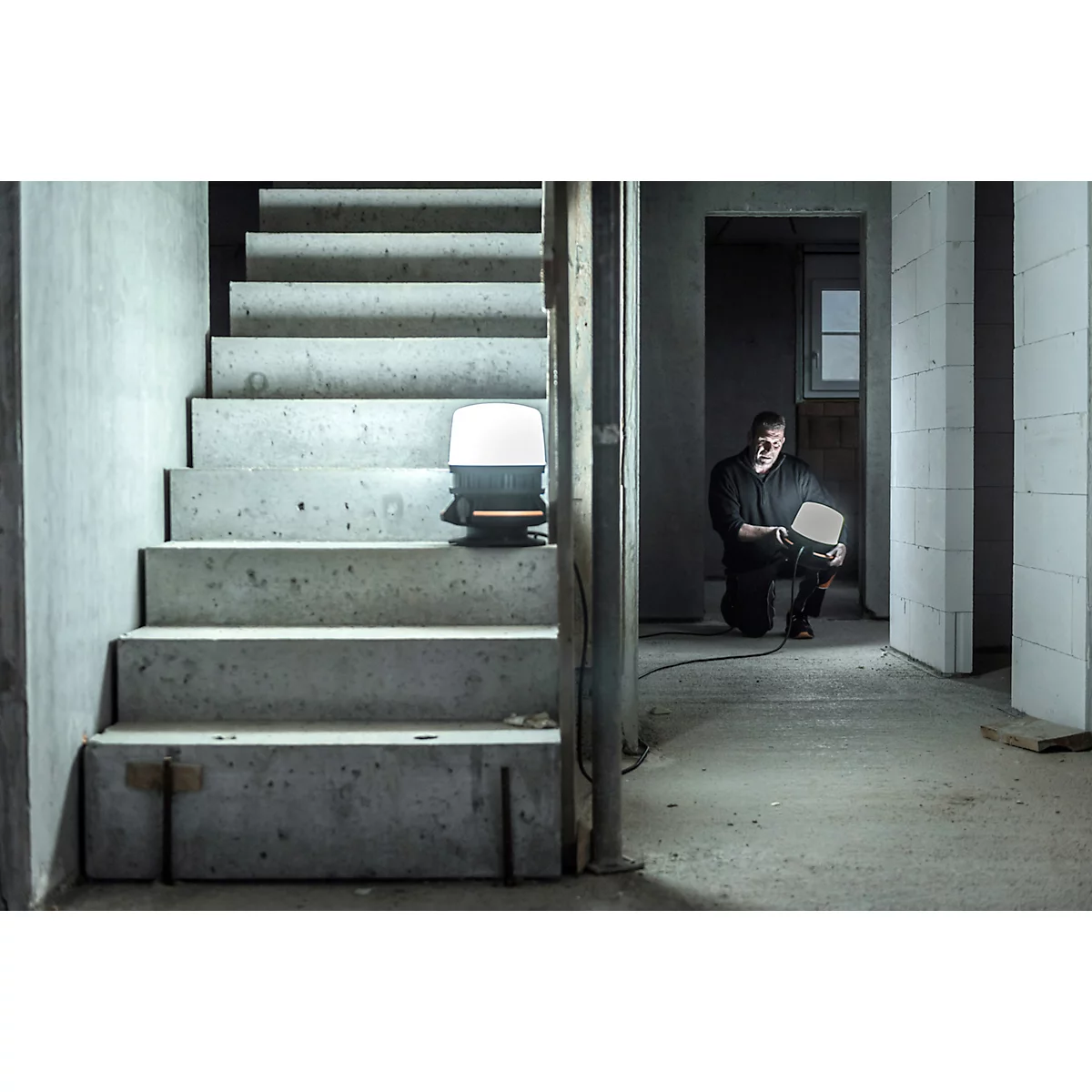 Un homme s'agenouille sur un chantier et tient une lampe de travail brillante. Escalier et lampe à droite, gros œuvre à gauche.