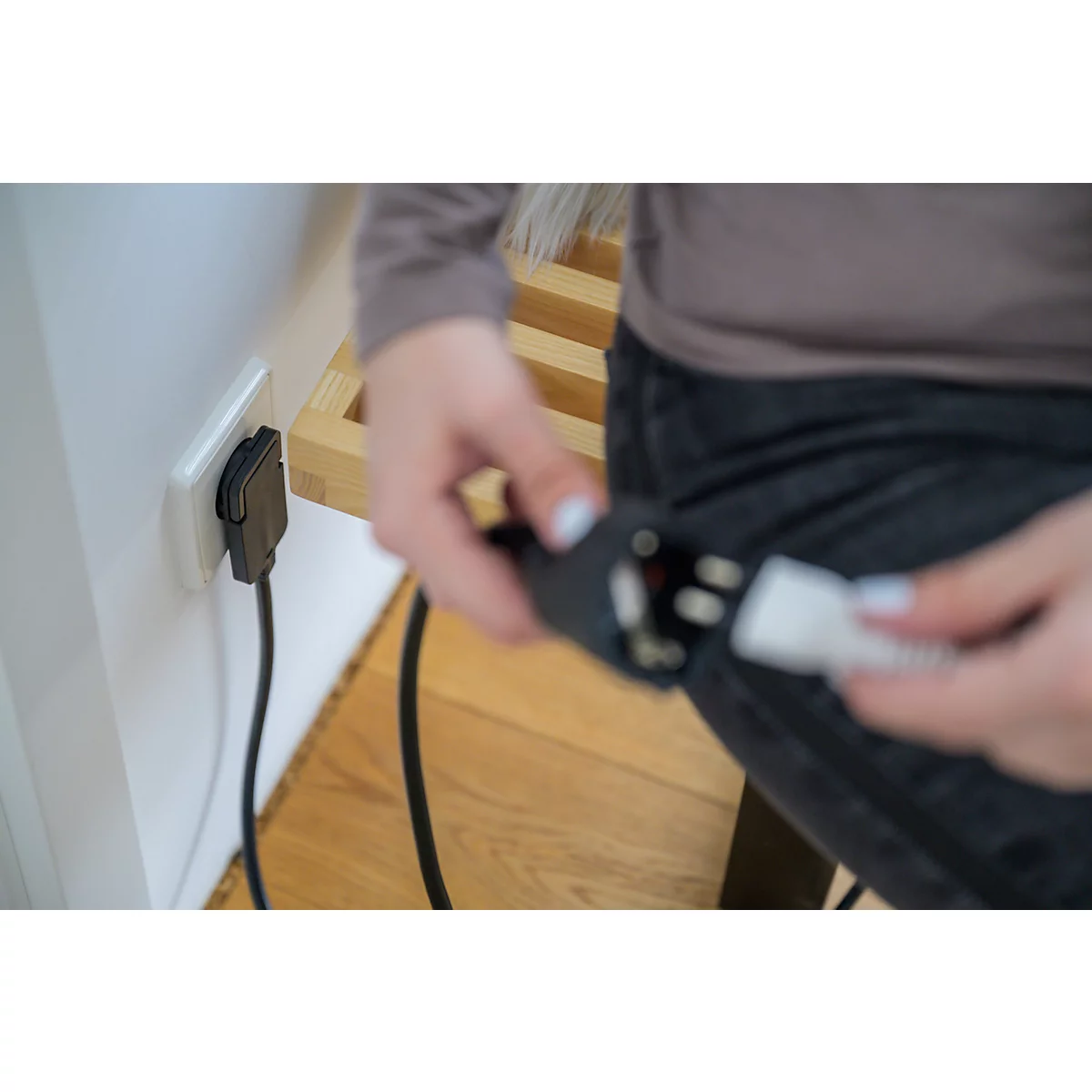 Un câble d'alimentation noir est branché dans une prise électrique blanche, sur un mur blanc. À droite, une étagère en bois clair.