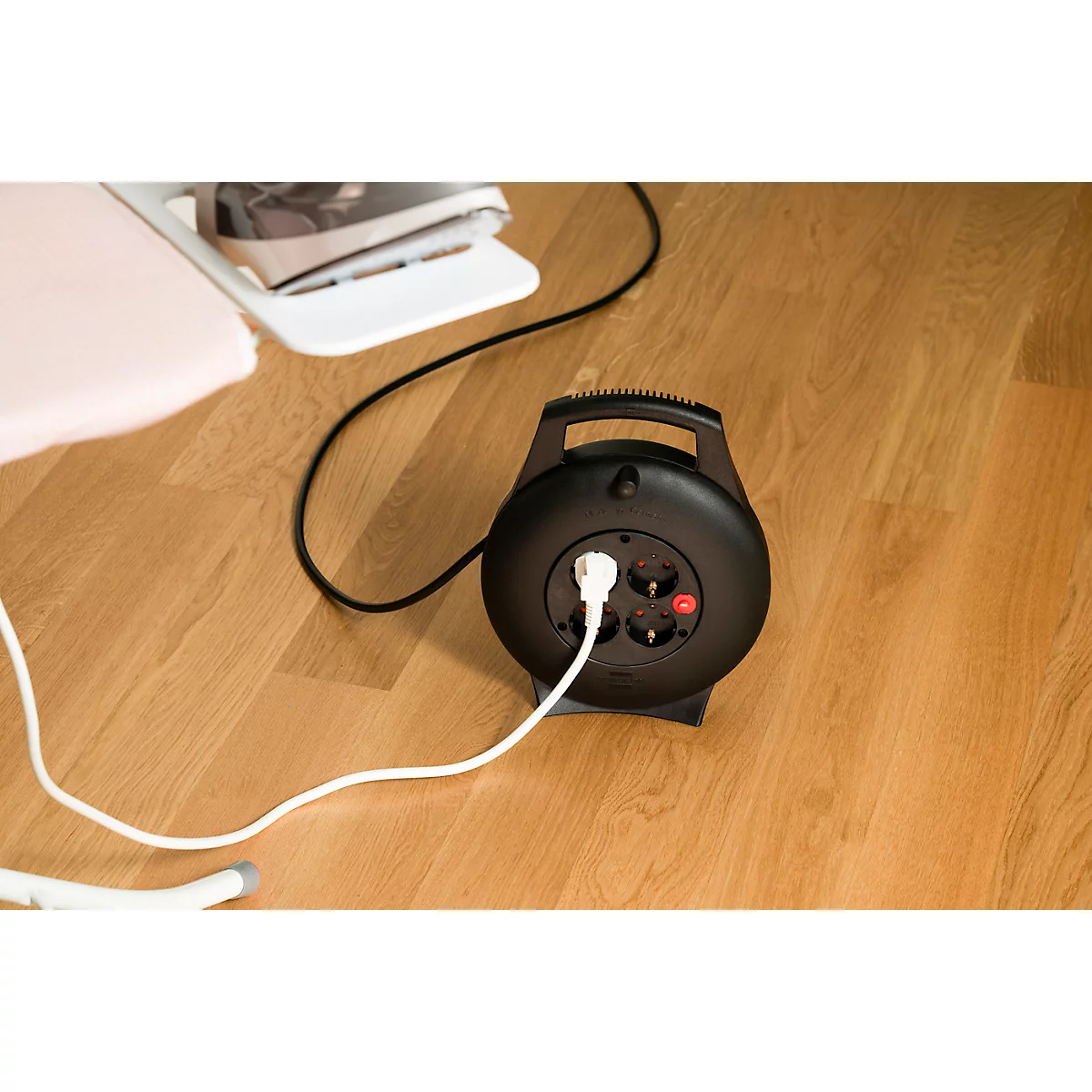 Schwarze Kabeltrommel mit eingestecktem weißen Stecker auf hellem Holzboden.