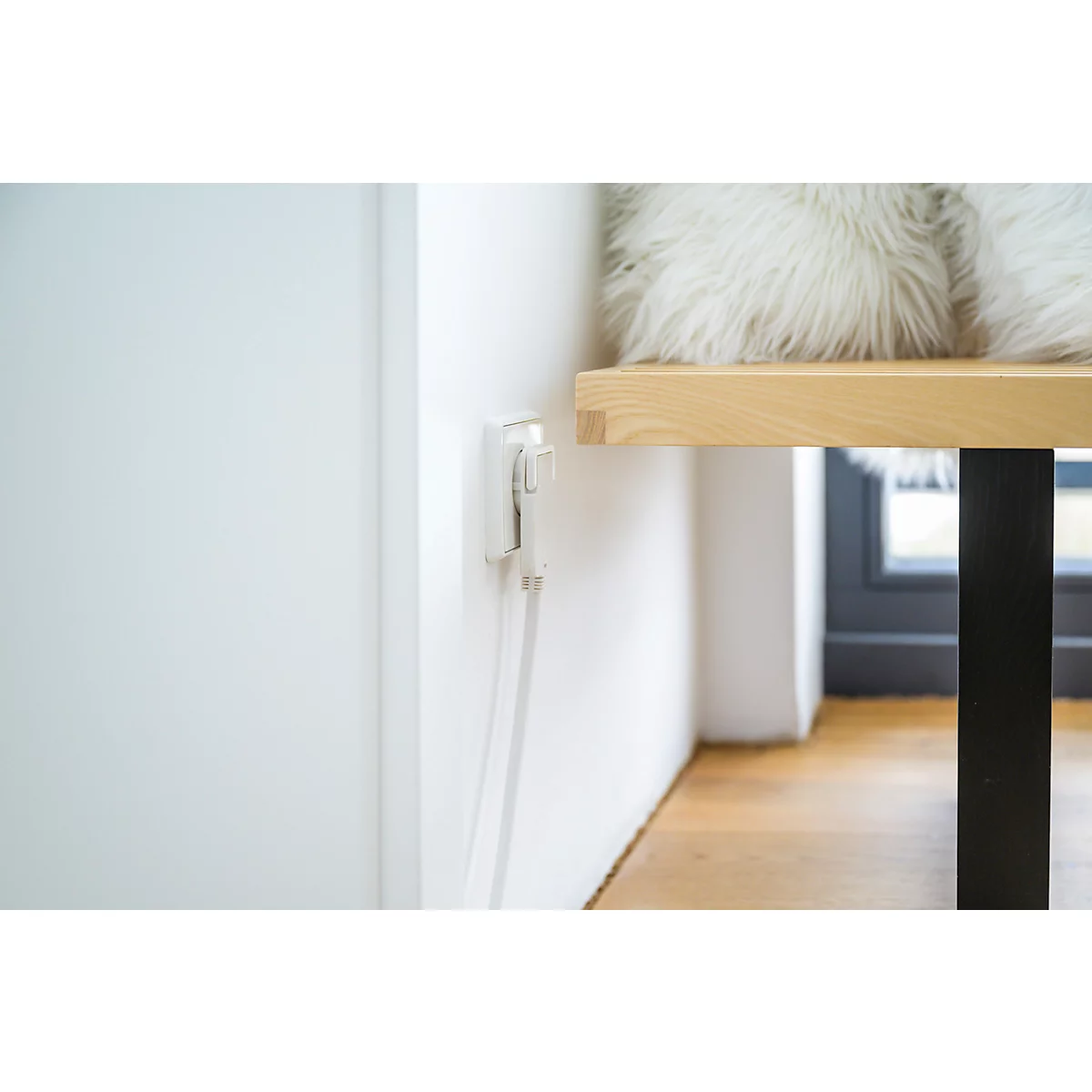 Close-up van een wit stopcontact aan een muur. Een witte kabel hangt naar beneden. Op de achtergrond: houten tafel met kussens.