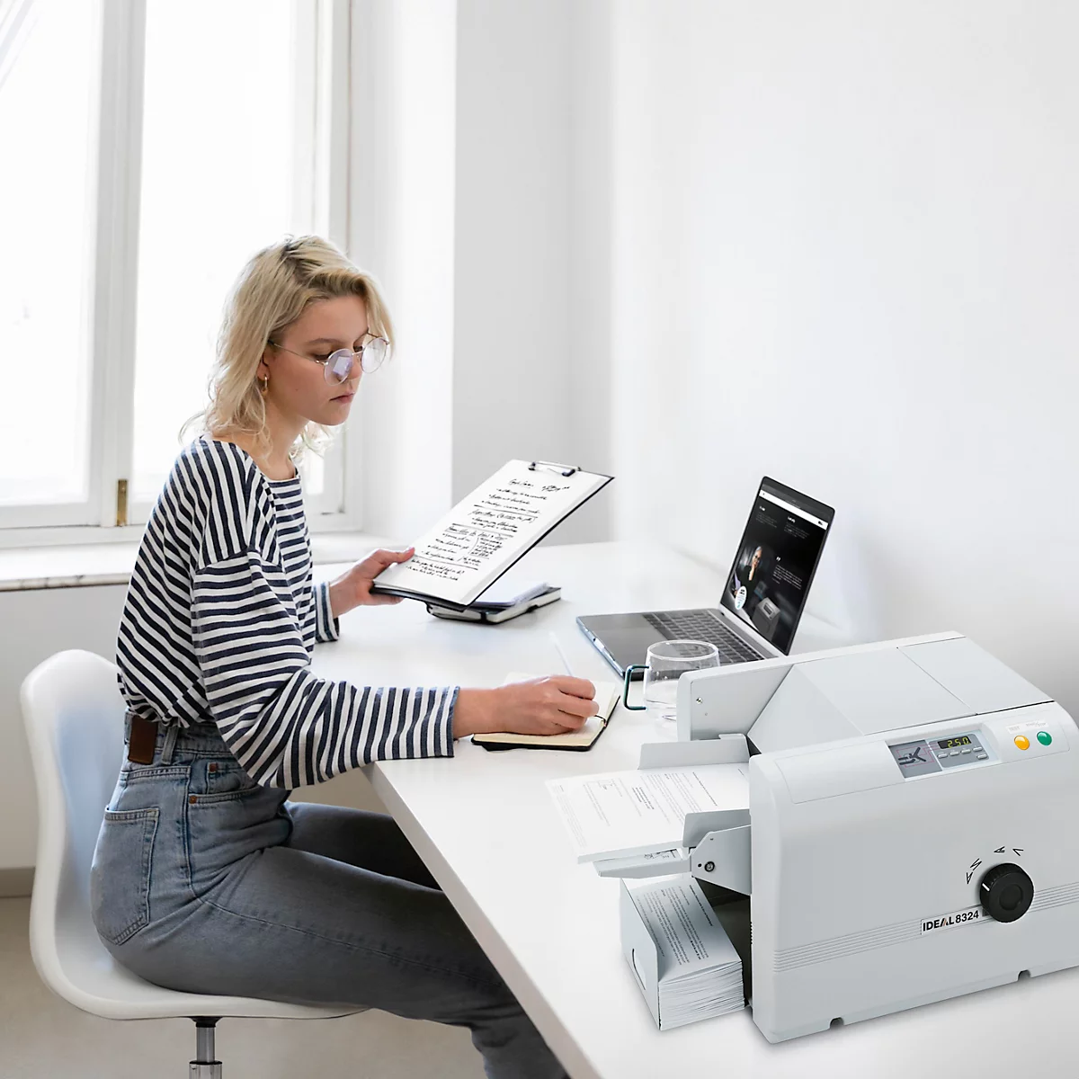 Frau sitzt an Schreibtisch, hält Klemmbrett und notiert etwas. Drucker und Laptop auf dem Tisch.