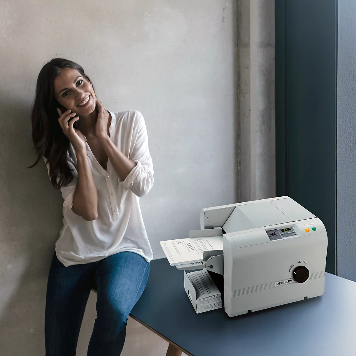 Eine lächelnde Frau telefoniert am Drucker. Der Drucker druckt Dokumente aus.