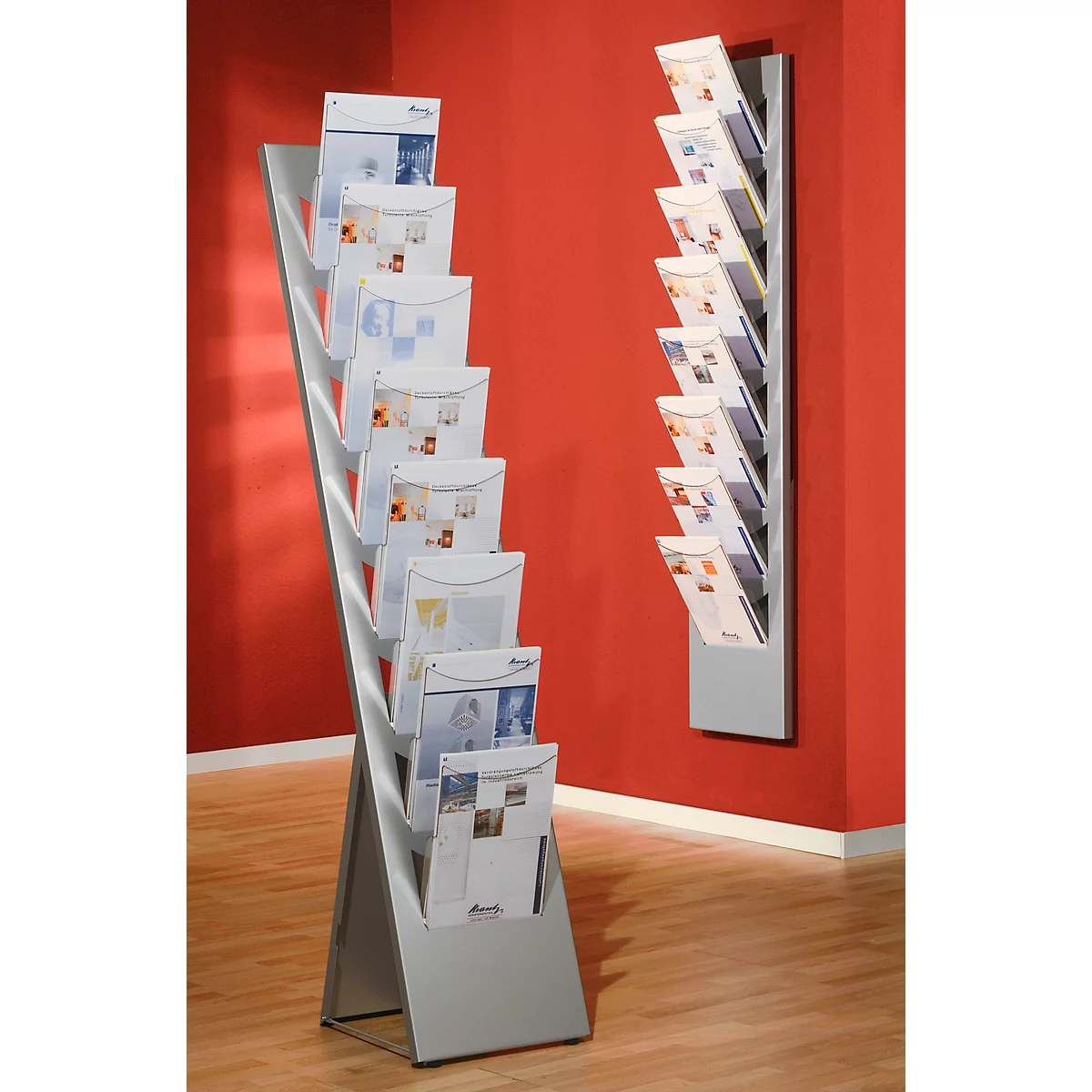 Two silver brochure stands with brochures in front of a red wall. The right stand is mounted on the wall.