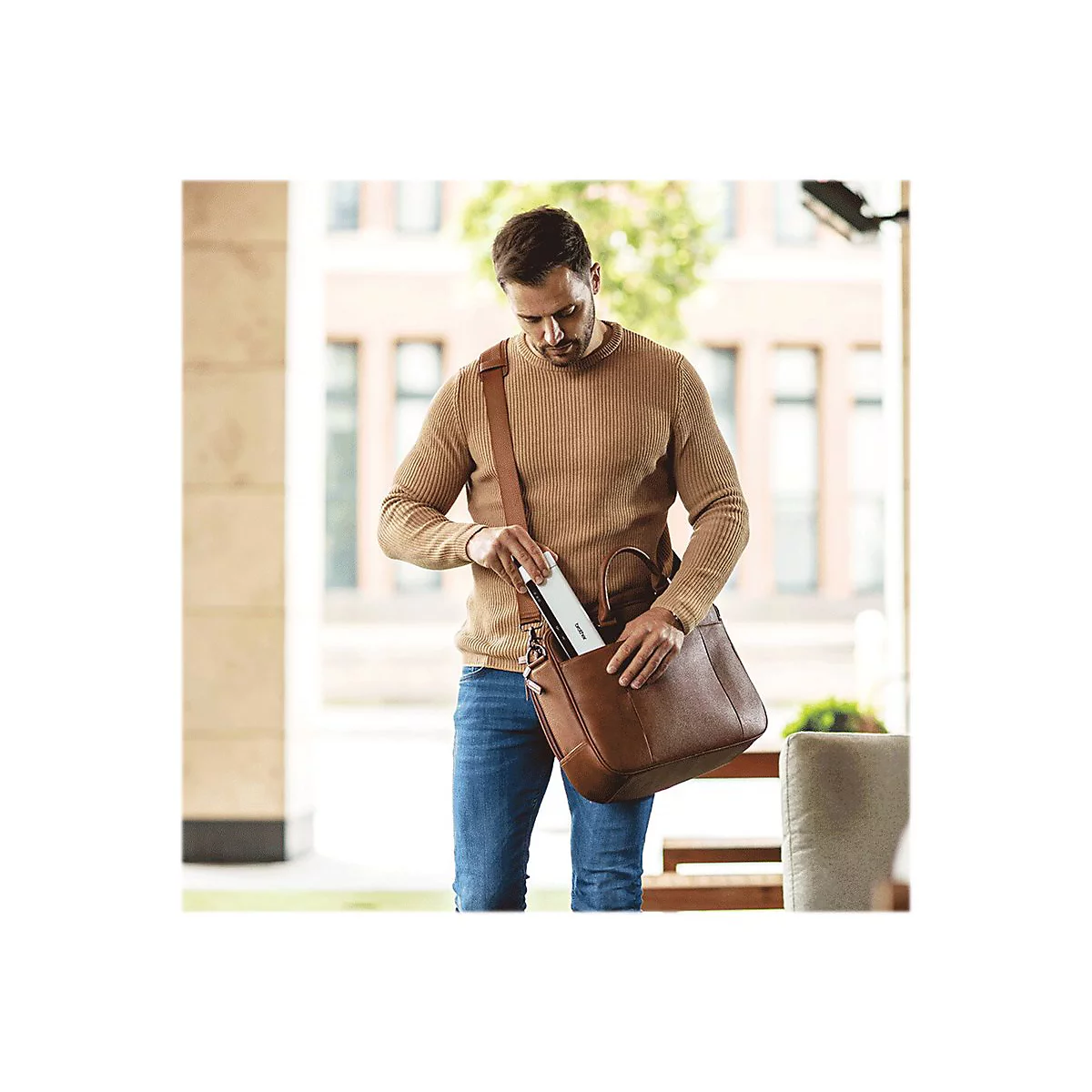 Ein Mann in Jeans und braunem Pullover holt ein weißes Gerät mit schwarzer Schrift aus seiner braunen Ledertasche.
