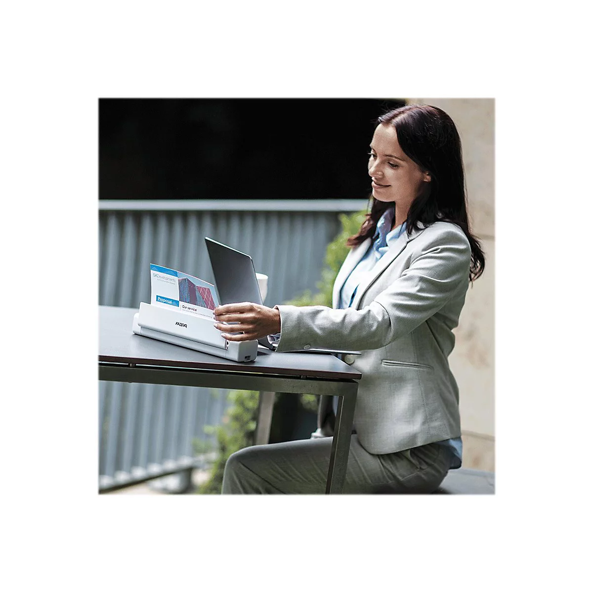 Eine Frau in einem grauen Blazer sitzt an einem Tisch und bedient einen mobilen Scanner. Auf dem Tisch liegt ein Laptop.