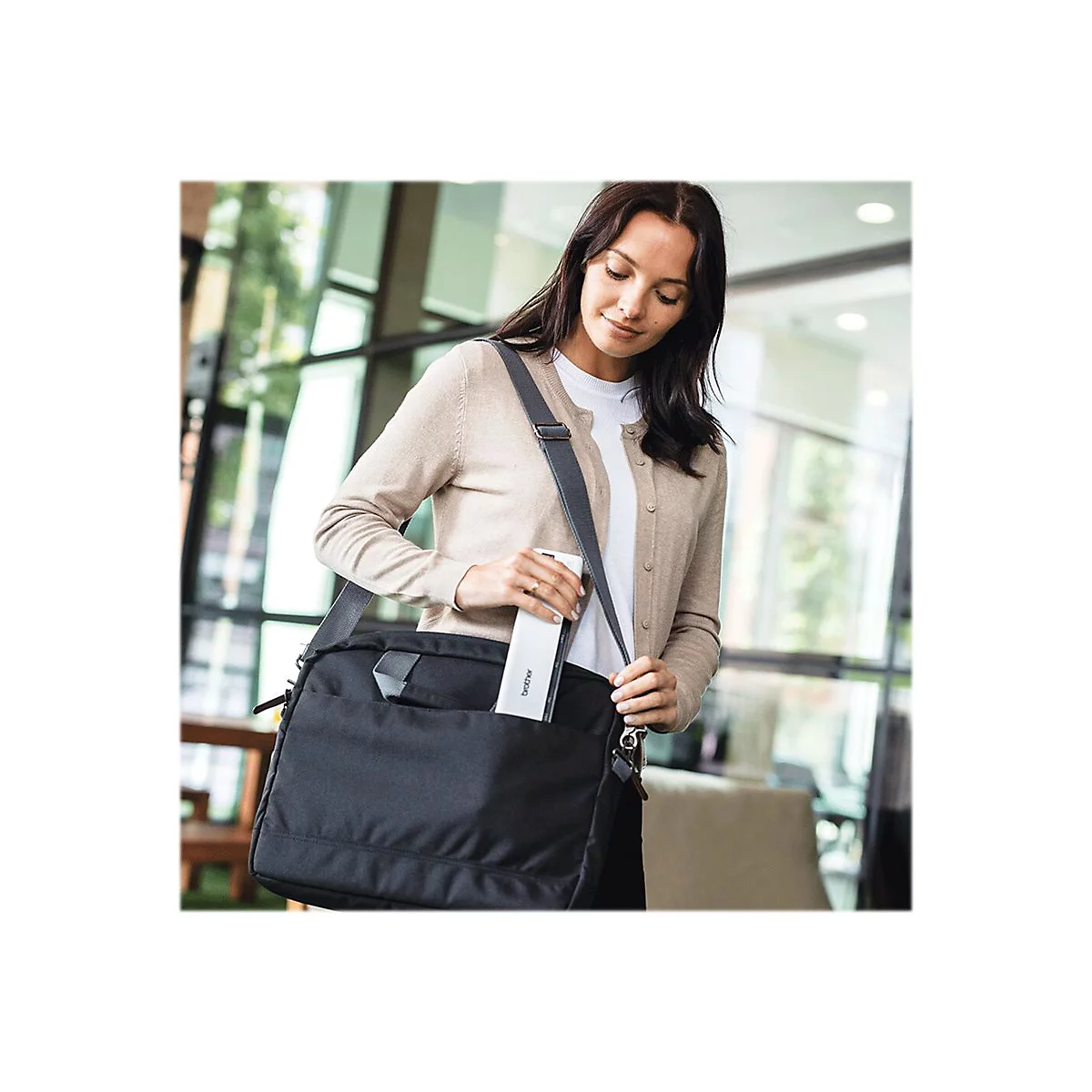 Eine Frau in hellbraunem Cardigan entnimmt einem schwarzen Messenger Bag ein Gerät. Im Hintergrund ist eine helle Büroumgebung zu sehen.