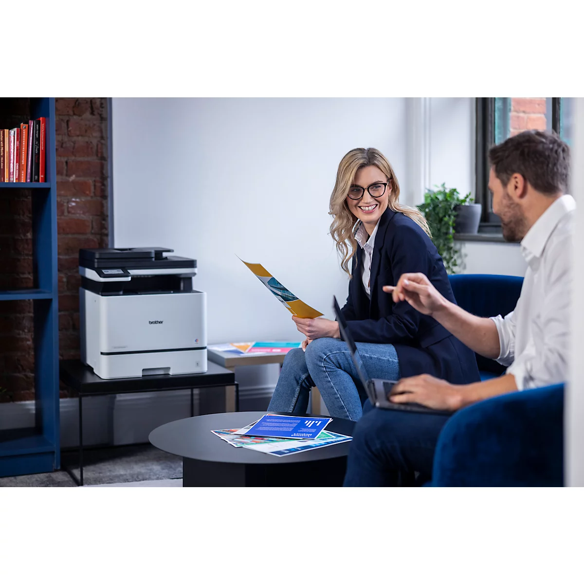 Eine lächelnde Frau und ein Mann arbeiten in einem modernen Büro zusammen, neben ihnen steht ein Brother-Drucker.