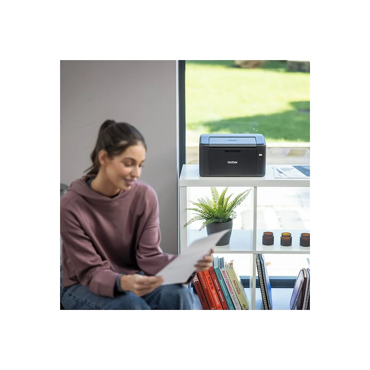 Eine Frau liest ein Dokument, während sie in der Nähe eines Druckers und eines Regals voller Bücher sitzt.