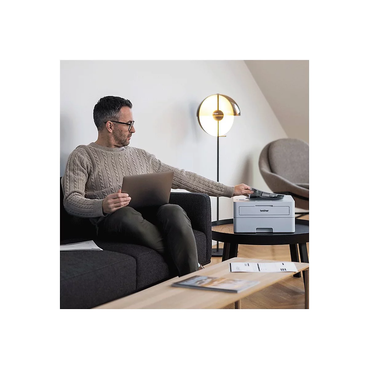 Ein Mann sitzt auf einem Sofa und bedient einen Drucker. Er hält einen Laptop und legt Papiere in den Drucker ein.