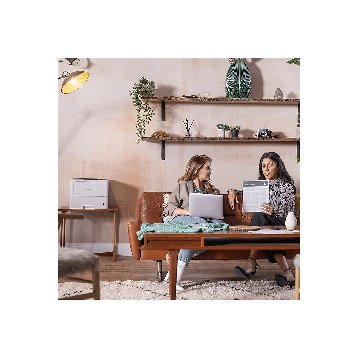 Zwei Frauen sitzen auf einem Sofa. Eine hält ein Dokument, die andere einen Laptop.