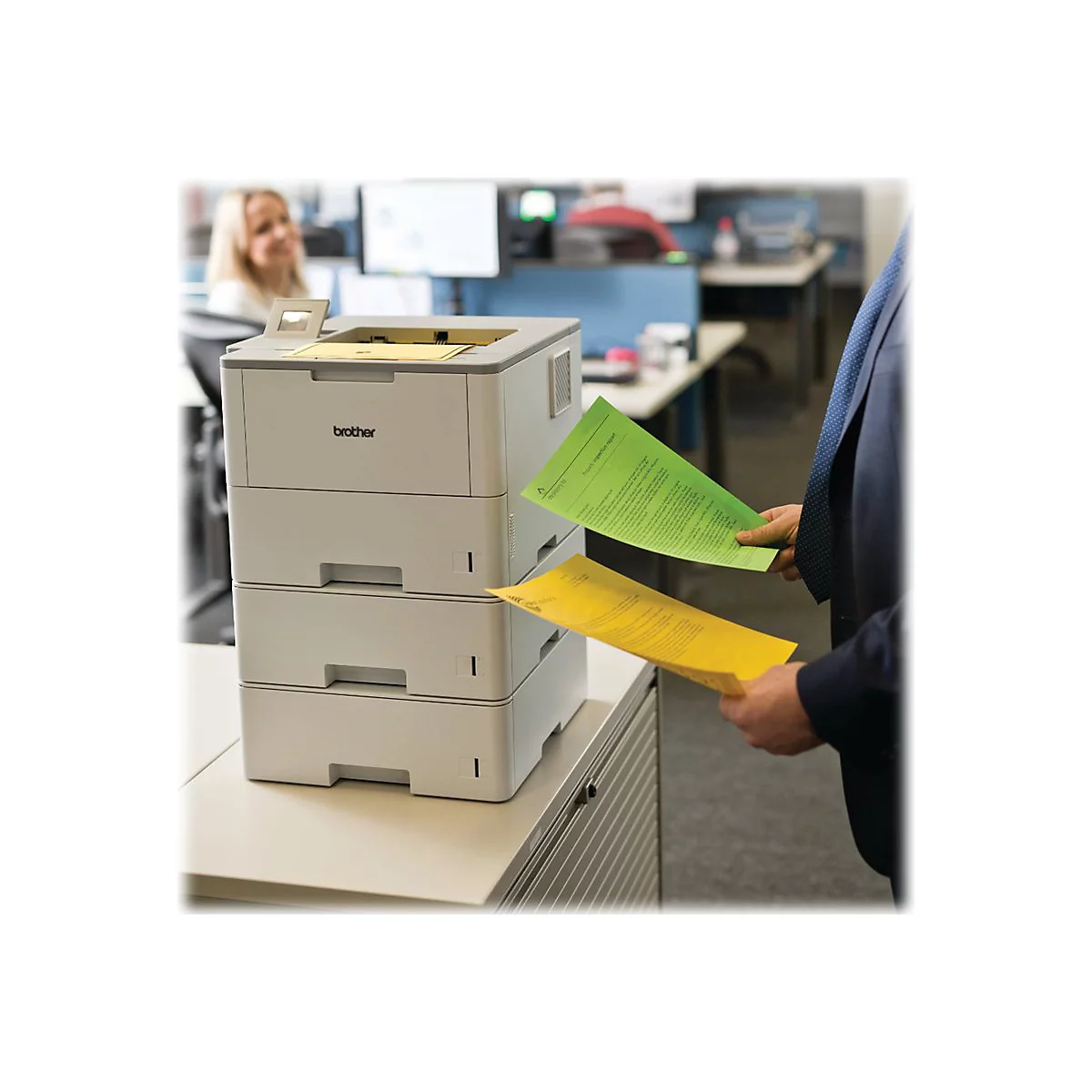 Ein Mann im Anzug hält zwei farbige Dokumente. Im Hintergrund ein Drucker und eine Frau.
