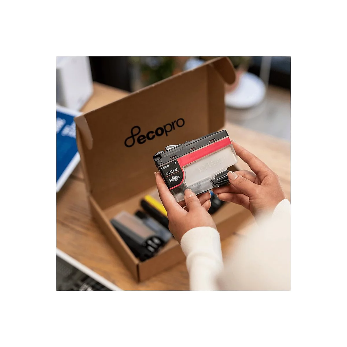 Hände halten eine Tintenpatrone mit rotem Streifen, im Hintergrund eine Schachtel und weitere Patronen.