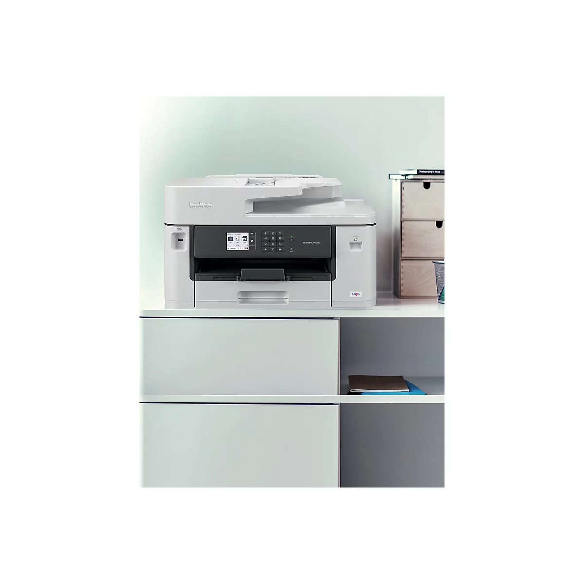 Ein weißer Multifunktionsdrucker steht auf einem Regal. Auf dem Display des Druckers werden Informationen angezeigt. Rechts davon ist ein Bücherstapel und Schubladen.