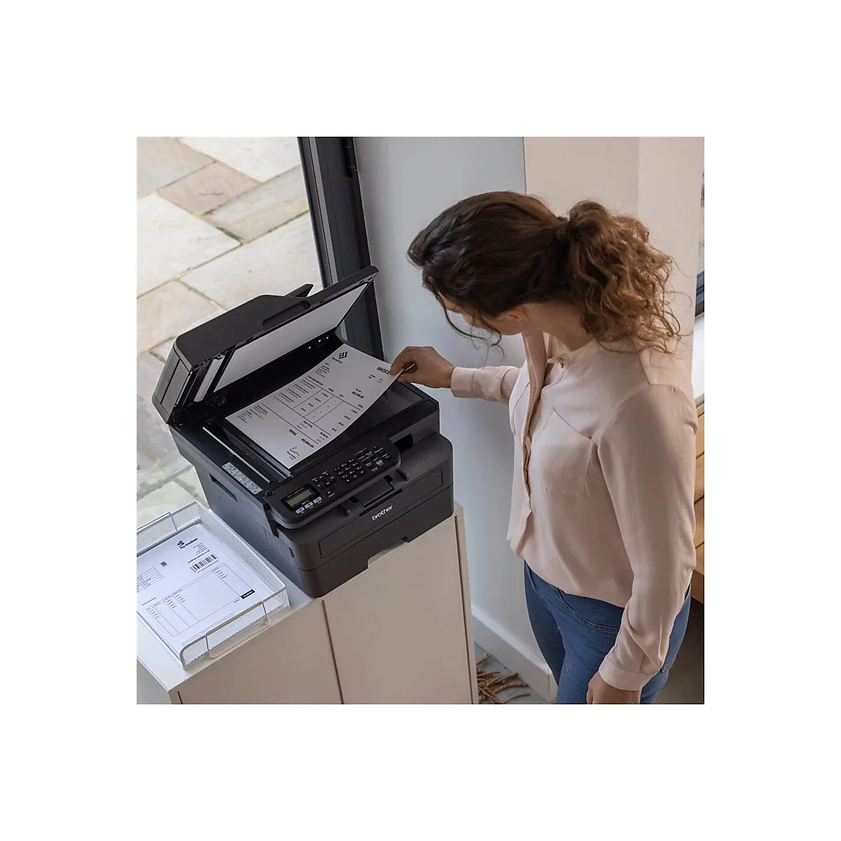 Eine Frau legt ein Dokument in einen schwarzen Drucker. In einer Ablage liegt ein gedrucktes Dokument. Der Drucker steht auf einem Schrank.