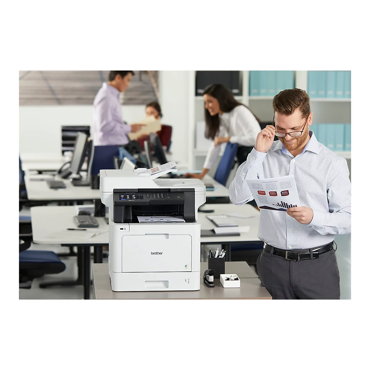 Ein Mann mit Brille betrachtet Dokumente neben einem Drucker. Im Hintergrund arbeiten weitere Personen.