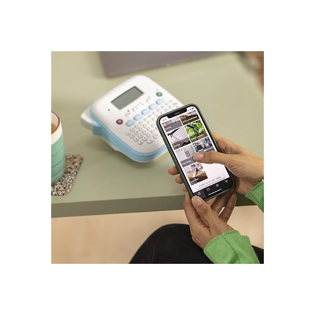 Eine Person hält ein Smartphone, auf dem eine App zum Etikettieren geöffnet ist. Ein Etikettiergerät liegt daneben. Auf dem Tisch sind eine Tasse und ein Untersetzer.