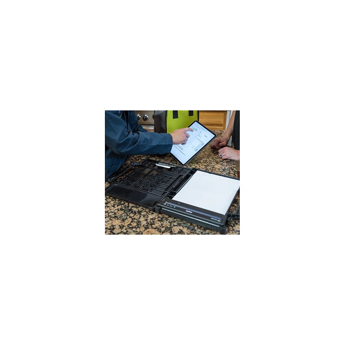 Person zeigt auf Tablet-Display mit Beleg. Vorne liegt ein Drucker mit Papier. Auf der ArbeitsflÃ¤che weitere Personen.
