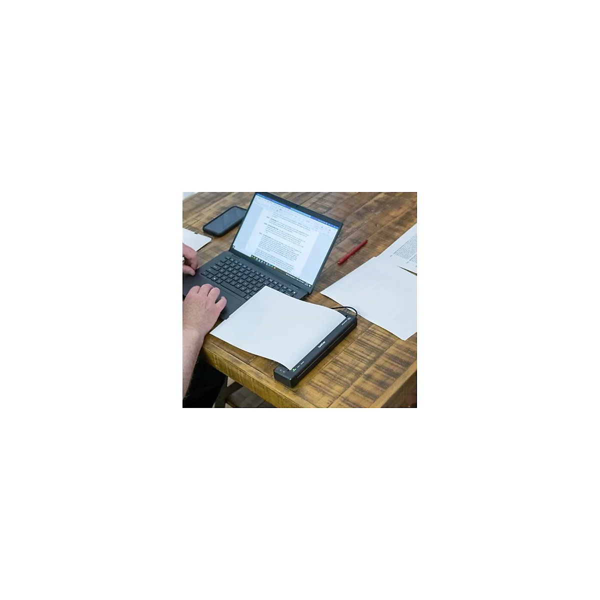 Person arbeitet an einem Laptop. Ein Blatt Papier liegt in einem Scanner. Auf dem Holztisch sind auch Smartphone und Stift.