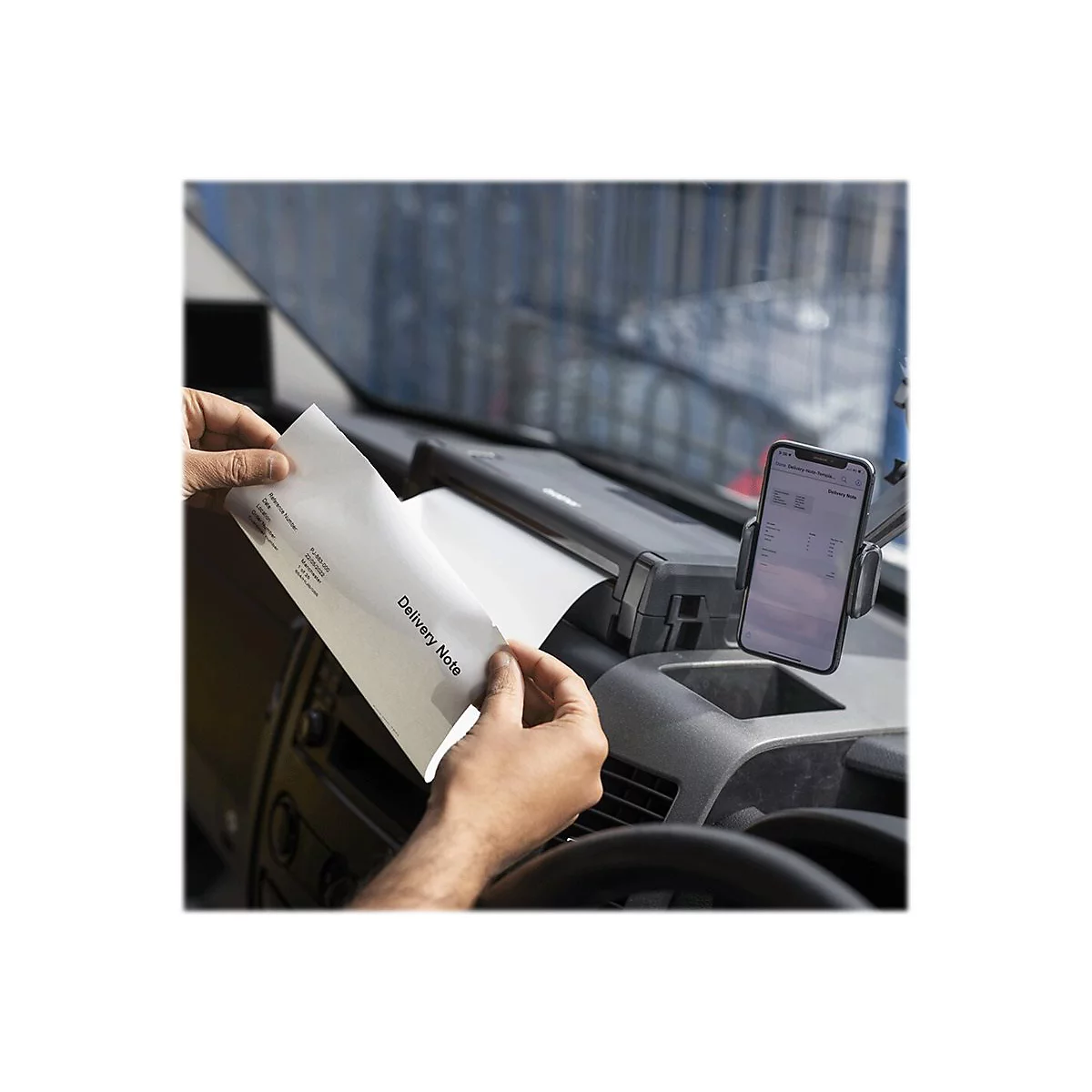 Person hält in einem Fahrzeug einen Lieferschein. Daneben ist ein Smartphone in einer Halterung zu sehen.
