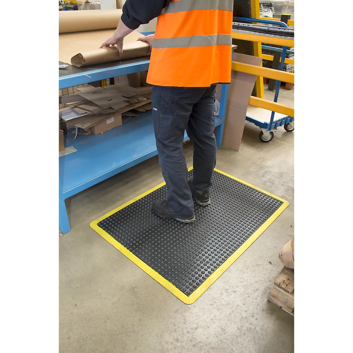 Person stands on a black mat with a yellow border in a factory. They are wearing a vest.
