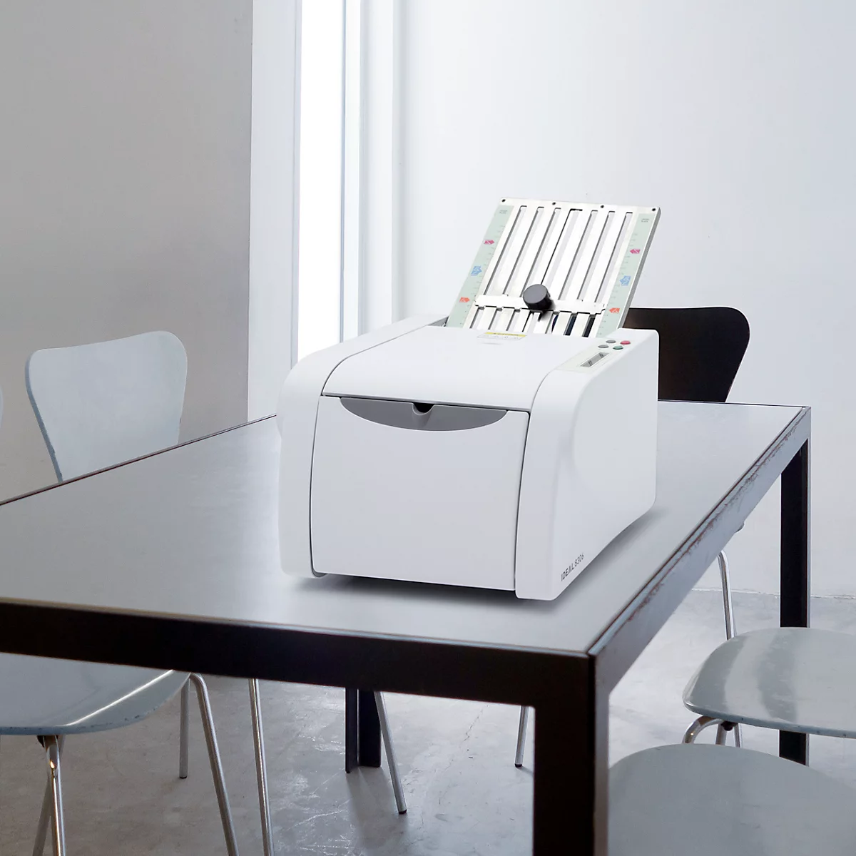 Weißer Drucker mit offenem Fach steht auf einem Tisch. Im Hintergrund Stühle, Fenster.