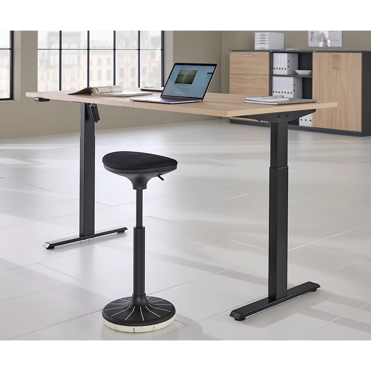 Een in hoogte verstelbaar bureau met een laptop en kruk in een modern kantoor. Een kast en een raam op de achtergrond.