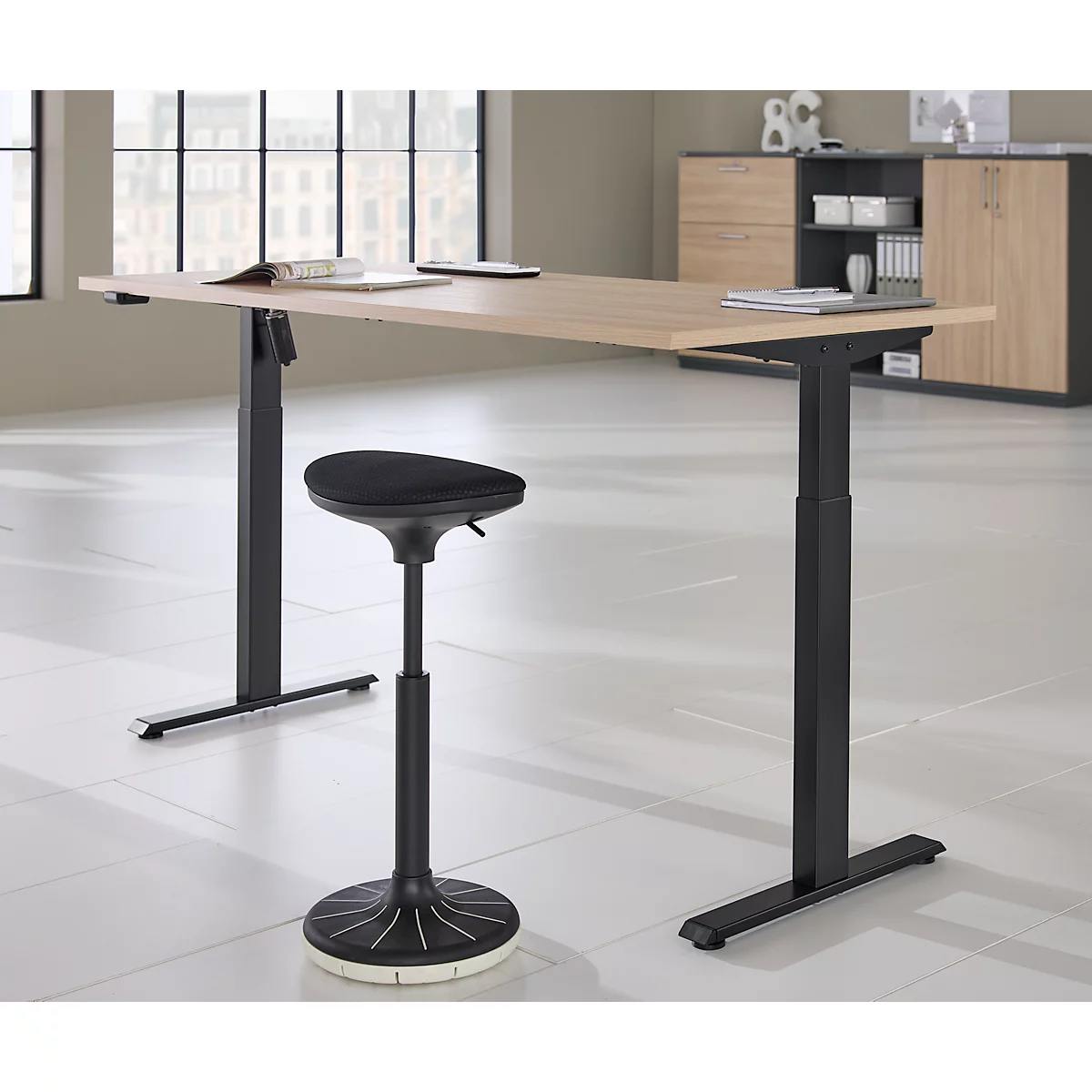Lichtbruine in hoogte verstelbare bureau met zwart frame, daarnaast een zwarte stahulp. Een kast op de achtergrond.