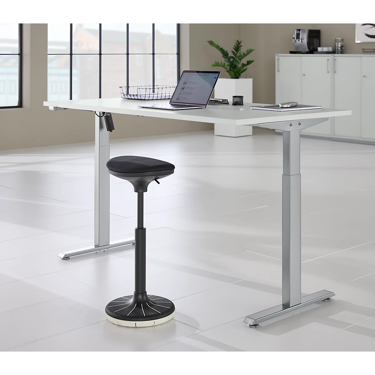 In hoogte verstelbaar bureau met laptop en kruk. Ramen en kasten op de achtergrond.