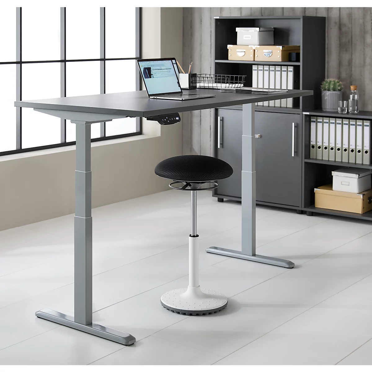 Bureau debout gris avec ordinateur portable, tabouret noir et armoire grise. L'arrière-plan est une fenêtre et un mur en bois.