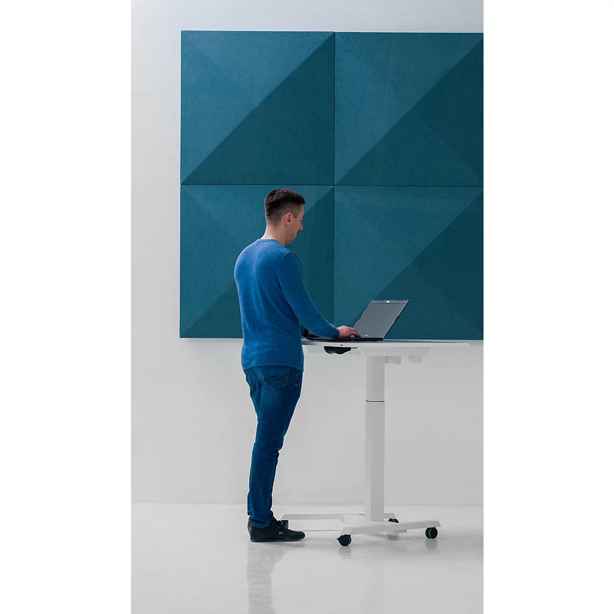 Un homme se tient devant un bureau blanc réglable en hauteur avec un ordinateur portable devant un mur bleu. Il travaille.