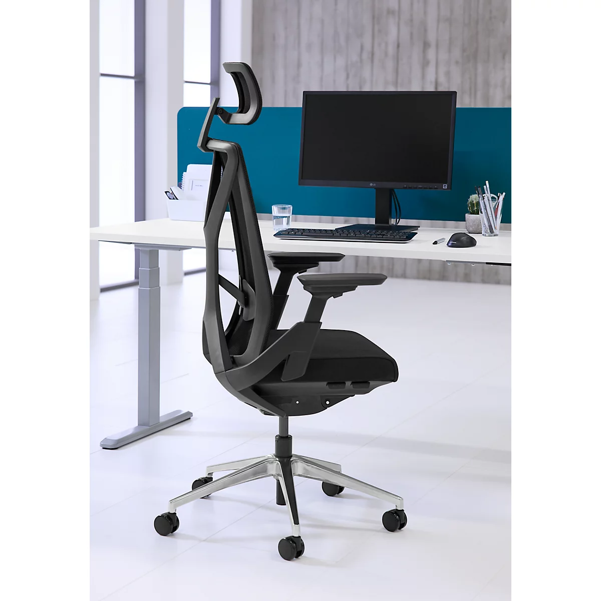Zwarte bureaustoel met mesh rugleuning voor een bureau met computer. Een blauw scheidingswand op de achtergrond.