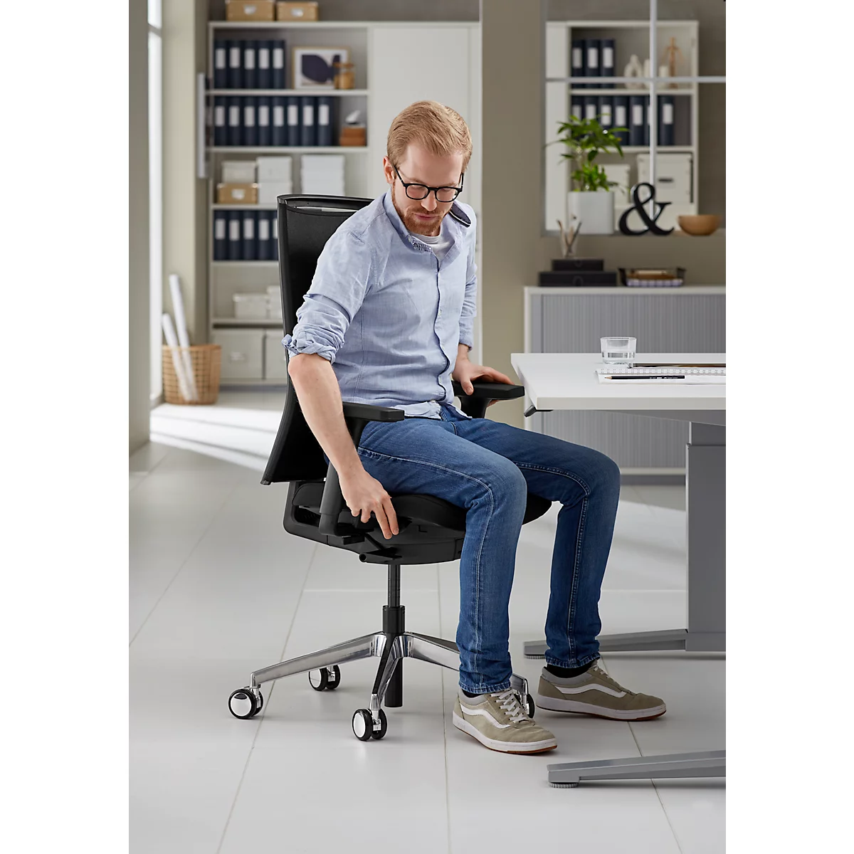 Een man op kantoor zit op een zwarte bureaustoel en stelt deze af. Hij draagt een spijkerbroek, een lichtblauw overhemd en een bril.