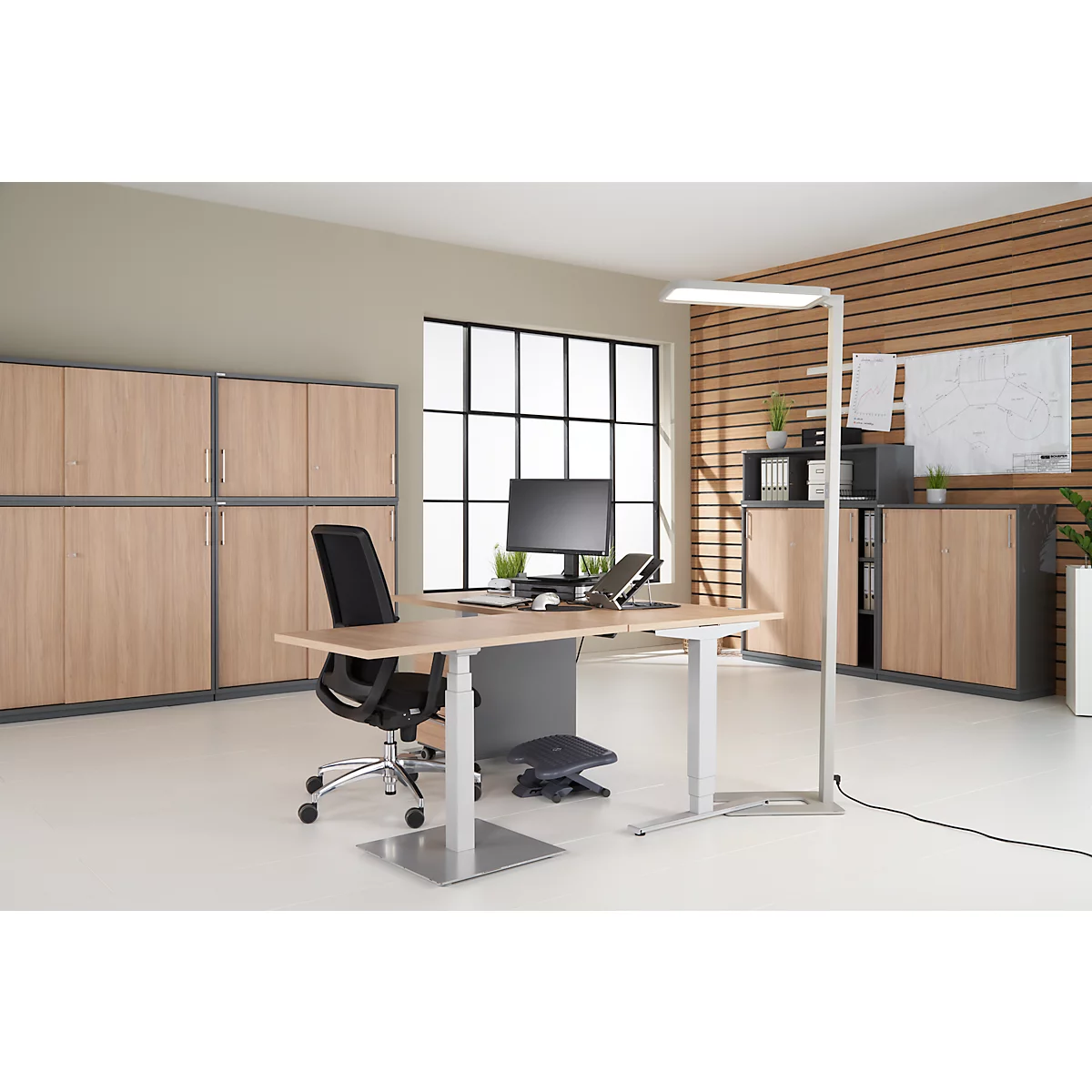 An office room with desk, chair, and cabinets. On the desk a computer, a lamp, and a footrest. Light wood and grey accents.