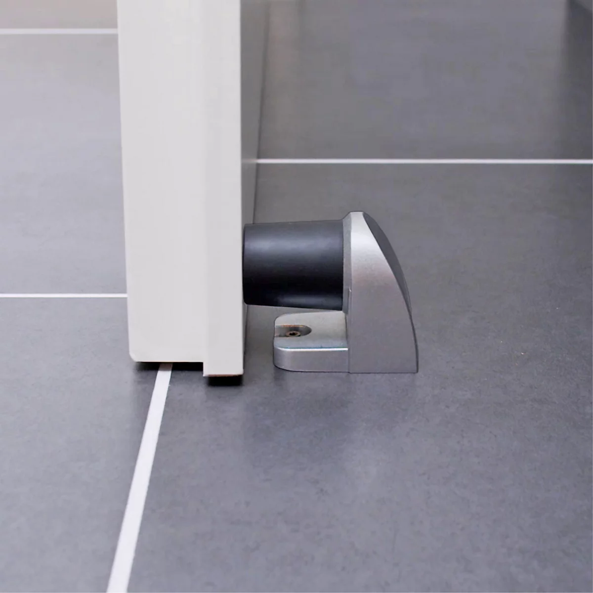 Un butoir de porte fixé à une porte blanche. Sur un sol gris. La lumière entre par le haut.